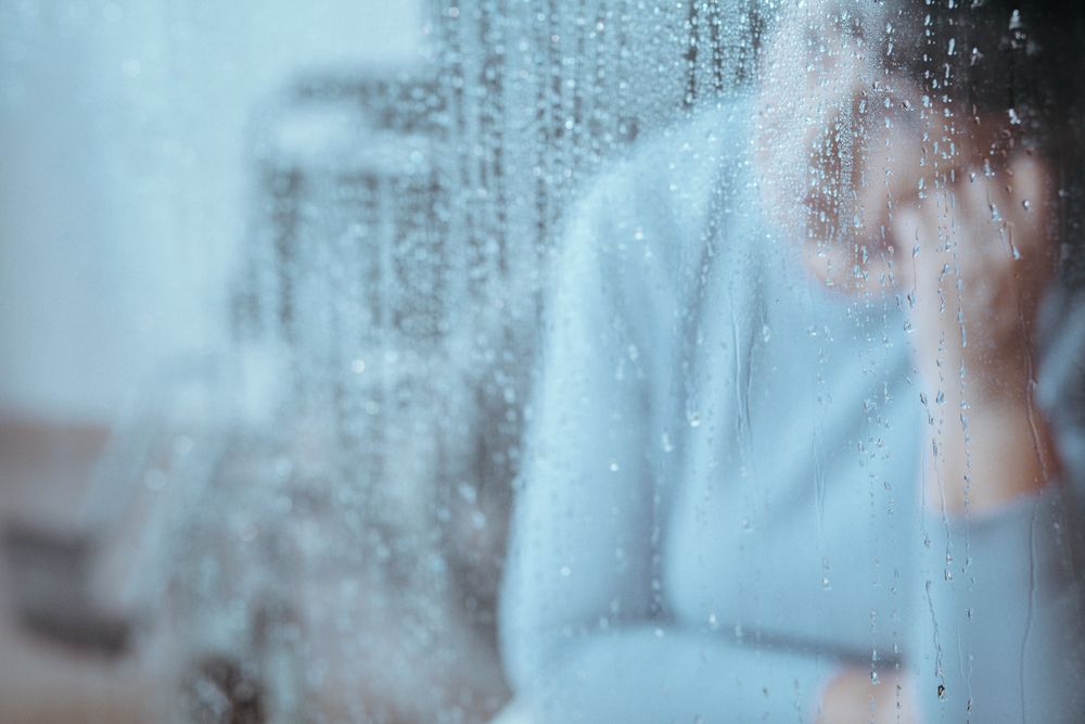 Blurred disabled senior woman with migraine behind window with raindrops