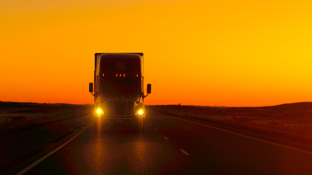 truck driver with a qualification file driving and hauling goods on empty highway across the Great Plains in golden morning.